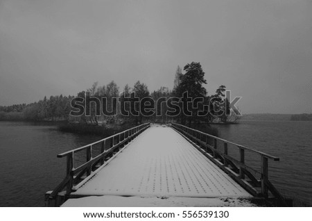 Snowy wooden bridge over lake in Sweden. Beautiful black and white nature and landscape photo. Cold cloudy day. Calm, peaceful photo. Nice outdoors image.