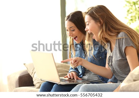 Two excited roommates reading good news on line with a laptop sitting on a couch in the living room at home with a window in the background Royalty-Free Stock Photo #556705432