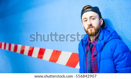 portrait of a young man near the blue wall color in the parking lot. A guy with a beard in a warm jacket posing on a parking