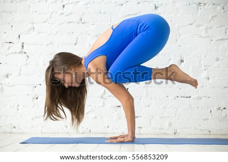 Young attractive happy smiling woman practicing yoga, standing in Crane exercise, Bakasana pose, working out, wearing sportswear, blue suit, indoor full length, white loft studio background