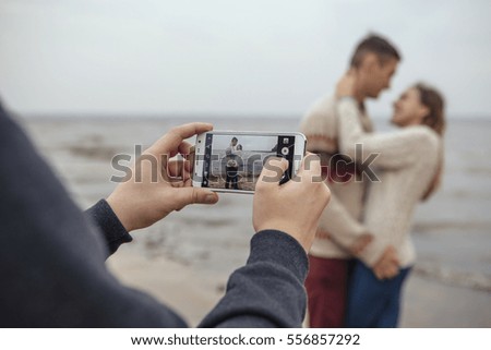 Happy thoughtful couple standing on a rock beach near sea hugging each other in cold foggy cloudy autumn weather. Man photograph them on a phone. Copy space