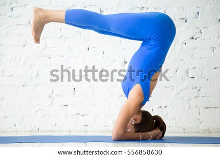 Young attractive woman practicing yoga, standing in variation of salamba sirsasana exercise, headstand pose, working out, wearing sportswear, blue suit, indoor full length, white studio background