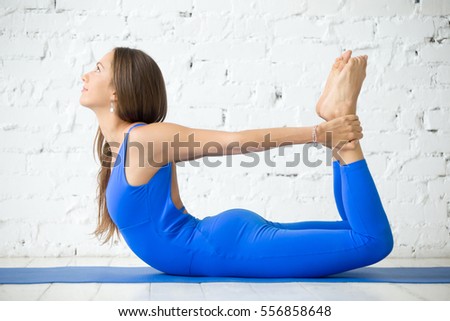 Young happy attractive woman practicing yoga, stretching in Bow exercise, Dhanurasana pose, working out, wearing sportswear, blue jumpsuit, indoor full length, white loft studio background