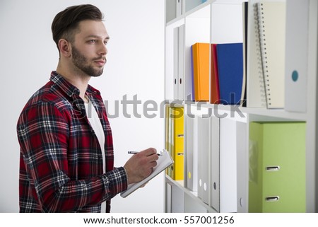 Side view of a serious bearded young man in a library. He is wearing a checkered shirt and taking notes
