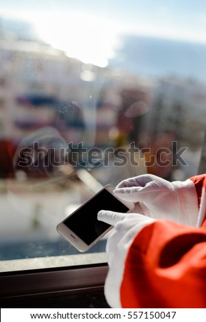 Santa Claus holding mobile smartphone in hands, mockup screen festive Christmas window background. Closeup view photo