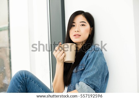 young beautiful asian woman drinking coffee or tea and looking outside on big modern windows