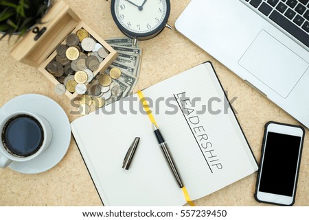 Modern wooden office desk table with laptop,smartphone, box of coins, paper money, table clock and cup of coffee. Writing Leadership on notebook page. Top view,flat lay