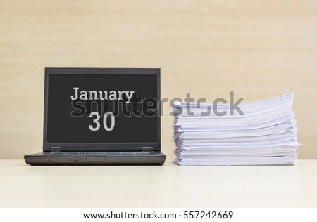 Closeup computer laptop with january 30 word on the center of screen in calendar concept and pile of work paper on wood desk and wood wall in work room textured background with copy space