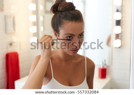 Young woman applying makeup at bathroom, brushing eyelashes.