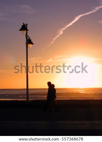 Backlit sunset on the beach with street lamp and people strolling
					