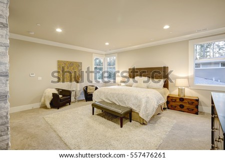 Beige and brown master bedroom boasts queen bed with taupe tufted headboard and bench. Beside the bed a vintage trunk nightstand next to sitting area with black leather armchairs. Northwest, USA
