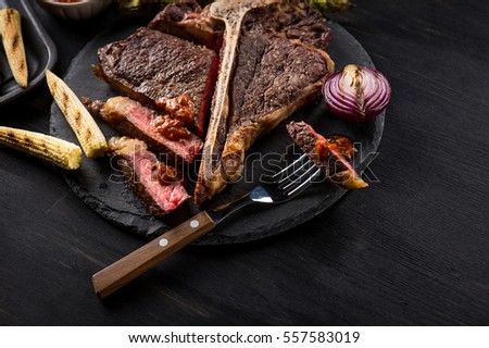 Sliced steak T-bone lying on a plate with grilled vegetables. Piece of steak on a fork. Black table.