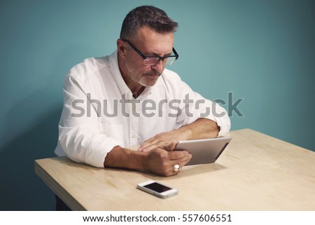 Portrait of concentrate male journalist in trendy eyewear reading book online on web page via modern touchpad using wireless connection during free time sitting near copy space for advertising