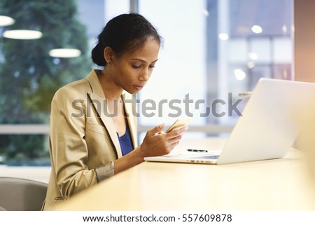 Concentrated female afro american journalist monitoring latest news in social networks via modern smartphone connected to wireless internet in office interior at the wooden table, copy space