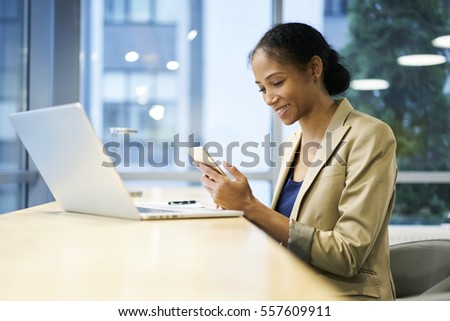 Smiling attractive afro american female downloading multimedia files from internet via modern smartphone connected to wireless network in office sitting near copy space area for advertising content