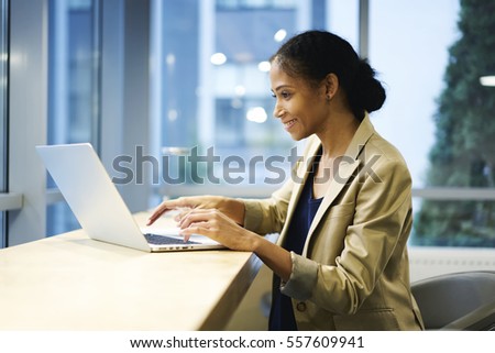 Talented afro American female editor of fashion magazine reading review and feedbacks on web page via modern laptop computer and wireless connection sitting on coworking copy space for advertising