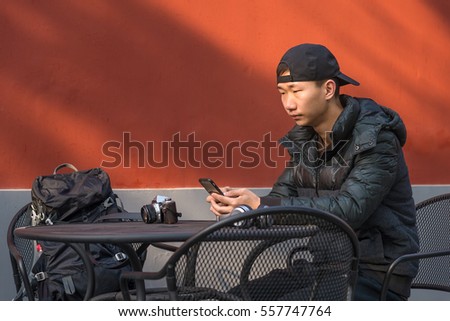 Asian handsome young man sitting besides coffee table holding a smart phone wit a Baseball cap in front of red wall