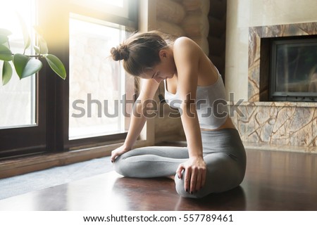 Young attractive woman practicing yoga, sitting in Uddiyana Bandha exercise, Upward Abdominal Lock pose, working out, wearing sportswear, grey pants, bra, indoor full length, home interior background Royalty-Free Stock Photo #557789461