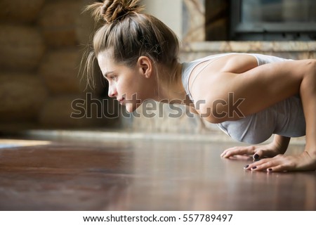 Closeup of young happy attractive woman practicing yoga, doing four limbed staff, push ups or press ups exercise, chaturanga dandasana pose, working out, wearing sportswear, bra, indoor, home interior Royalty-Free Stock Photo #557789497