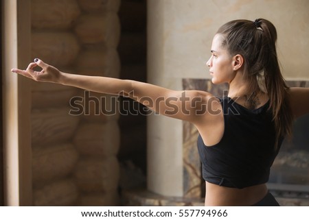 Young attractive woman practicing yoga, standing in Warrior Two exercise, Virabhadrasana 2 pose, working out, wearing sportswear, black tank top, shorts, indoor full length, home interior