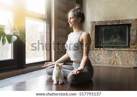 Young attractive smiling woman practicing yoga, sitting in Half Lotus exercise, Ardha Padmasana pose, working out, wearing sportswear, meditation session, indoor full length, home interior, cat near Royalty-Free Stock Photo #557795137