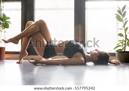 Young attractive woman practicing yoga, stretching in Fish exercise, Matsyasana pose, working out, wearing sportswear, black tank top, shorts, indoor full length, home interior background