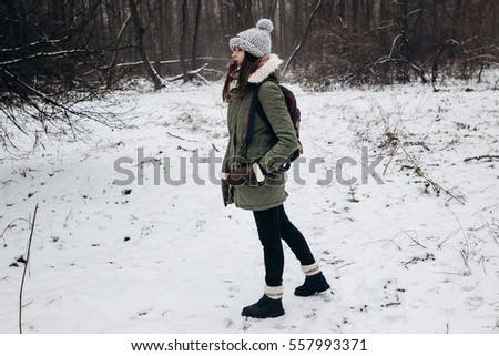 stylish hipster traveler woman with backpack walking in winter snowy forest trees with photo camera. wanderlust and adventure concept with space for text. atmospheric moment 