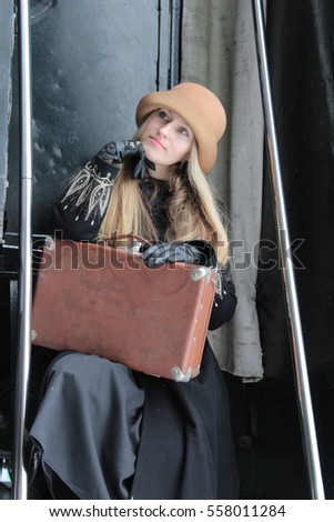 Portrait of a beautiful young woman. Vintage hat, old suitcase. Retro train. Travel concept. Winter coat, gloves. Vintage classic clothing. Blond girl. Reproduction and staging.