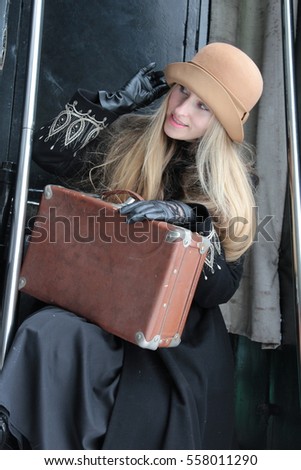 Portrait of a beautiful young woman. Vintage hat, old suitcase. Retro train. Travel concept. Winter coat, gloves. Vintage classic clothing. Blond girl. Reproduction and staging.