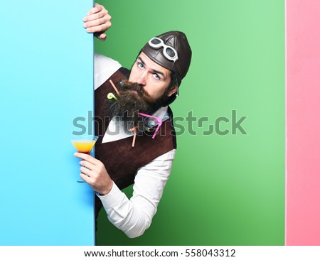 handsome bearded pilot or aviator man with long beard and mustache on serious face holding glass of alcoholic beverage in vintage suede leather waistcoat with hat and glasses on studio background