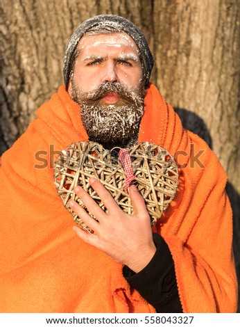Frozen man with squinty eyes , bearded hipster, with beard and moustache covered with white frost wrapped in orange blanket with wicker heart on winter day outdoors on natural background