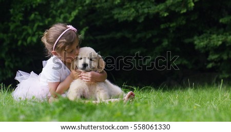 Cute toddler little two years old girl gives a kiss to a golden retriever puppy on a green widow in a woods. Concept of love for nature, protection of animals,innocence, fun, joy, carefree childhood.