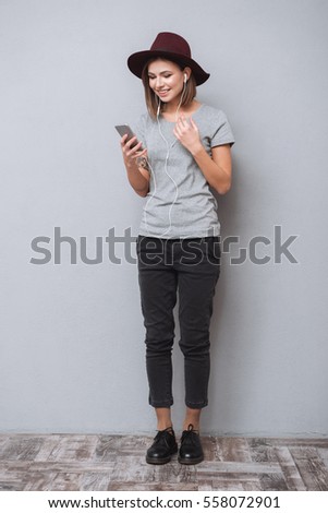 Full length portrait of a smiling young woman in hat standing and listening music with earphones isolated on a gray background