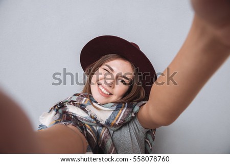 Portrait of a laughing woman in hat and scarf making selfie photo and winking over gray background