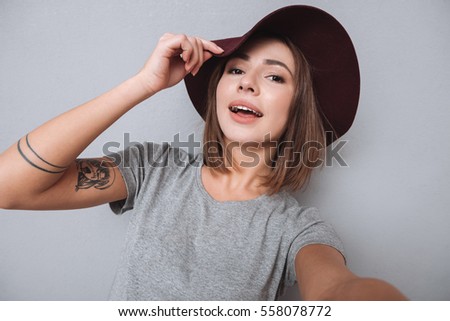 Portrait of an attractive young girl in hat taking selfie photo over gray background