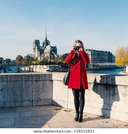 Young blonde woman full body portrait taking photo with camera with Notre Dame Cathedral in the background. Paris, France. 