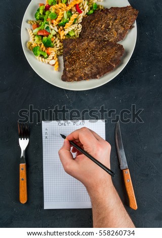 Concept of dieting, counting calories: a plate of healthy food (whole grain pasta with vegetables); man fills out a food diary. Hands in picture, top view, copy space 