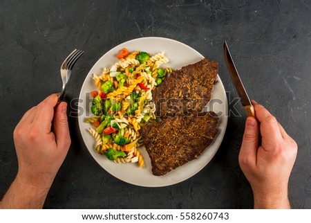 Concept of healthy way of life, my plate: a portion of whole grain pasta with vegetables and beef steak. Top view, man's hands with fork and knife in the picture 