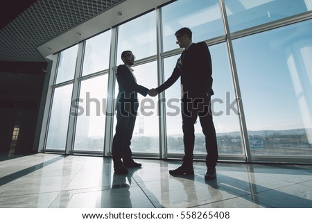 Two young businessmen are shaking hands with each other standing against panoramic windows. Royalty-Free Stock Photo #558265408