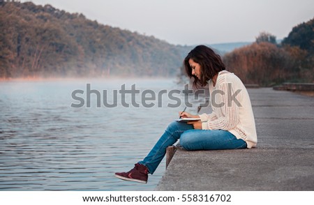 Girl writes diary by lake Royalty-Free Stock Photo #558316702