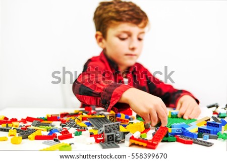 Distance learning. Home education. Blurred happy boy eight years, playing game of small plastic toy building blocks on white table on white background                                