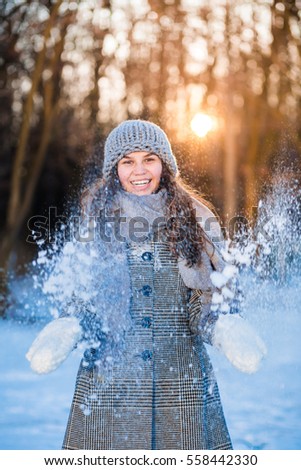 Joyful beauty girl in gray hat blowing snow in frosty winter park. Sunny day. 