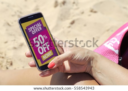 Young woman resting on the beach in the sun with a smartphone where she reads 50% off advertising on the screen.  Marketing, ecommerce, cell phone publicity.