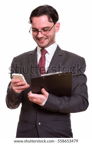 Studio shot of young happy businessman smiling while using mobile phone and holding clipboard isolated against white background