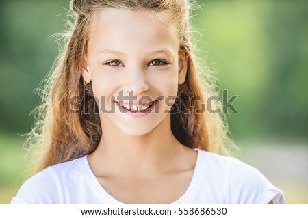 Portrait of beautiful smiling teenage girl in white blouse, against green of summer park.
