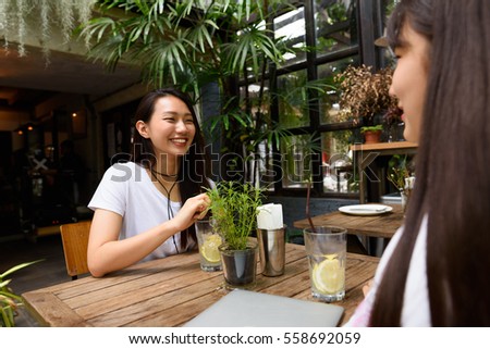Two happy teenage Korean friends smiling and sitting together with lemon iced tea outside nature themed restaurant