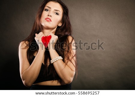 Woman brunette long hair girl wearing black dress holding red heart love symbol studio shot on dark. Heartbroken young female. Sad unhappy face expression