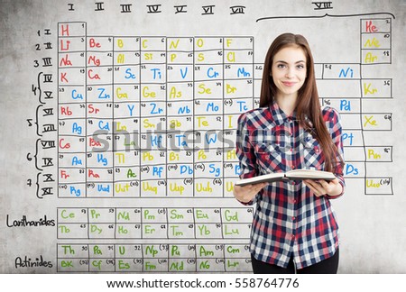 Close up of a nerdy teen girl in a checkered shirt holding an open book near a concrete wall with Mendeleev periodic table on it.
