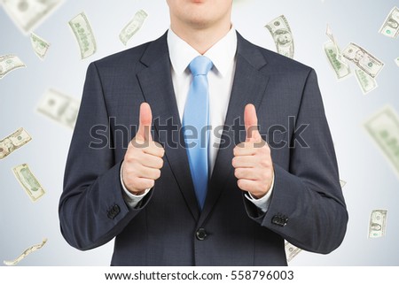 Close up of a businessman in a suit wearing a blue tie. He is showing thumbs up and standing against a gray wall with dollar bills falling.