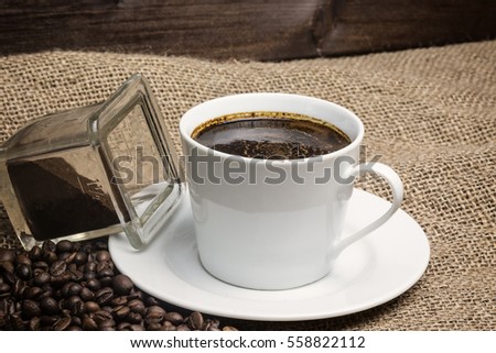 Closeup view of the vintage photo of the white cup of coffee standing on the sackcloth. Around it are scattered coffee beans and ground coffee glass  